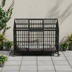 Black metal dog crate with wheels on a tiled floor, surrounded by potted plants.